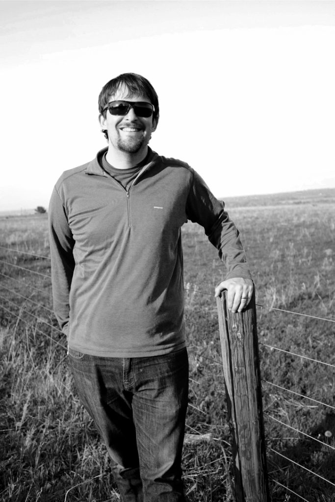Black and white photo of Blake Reid, a white man with short, dark hair and a goatee, wearing jeans, a T-shirt, a pullover, and sunglasses, leaning against a post with grass in the background