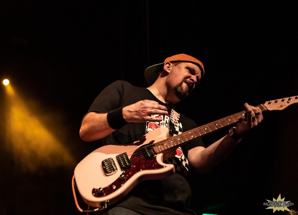 Photo of Blake Reid, a white man with a goatee, playing a pink G&L Fallout guitar with open A chord fretted, a checkerboard strap with a pink lightning bolt, wearing a black wristband, a black shirt with an obscured 12 Cents for Marvin Santa logo, a spotlight shining through haze in the background, and the logo of Backstage Flash 